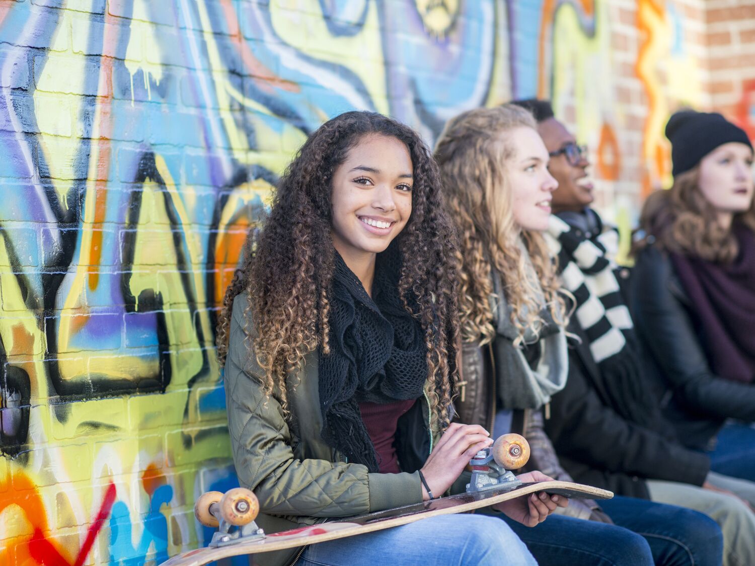 A high school student of African descent is hanging out with her friends. She is holding a skateboard and smiling at the camera.