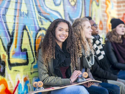 A high school student of African descent is hanging out with her friends. She is holding a skateboard and smiling at the camera.