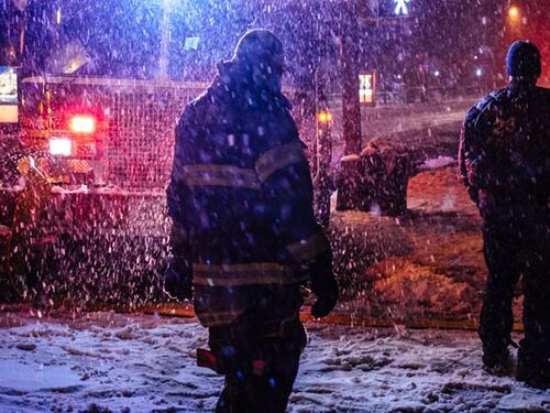 Firefighters standing in heavy snowfall at night, illuminated by emergency vehicle lights as they work at a snowy scene.