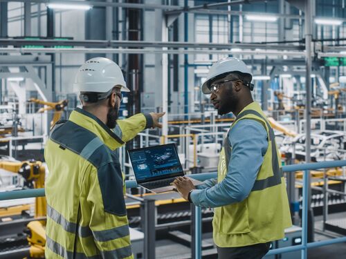 Two Car Factory Specialists in High Visibility Vests Using Laptop Computer. Engineers Discussing Automotive Industrial Manufacturing Facility Process of Vehicle Production. Automated Assembly Plant.