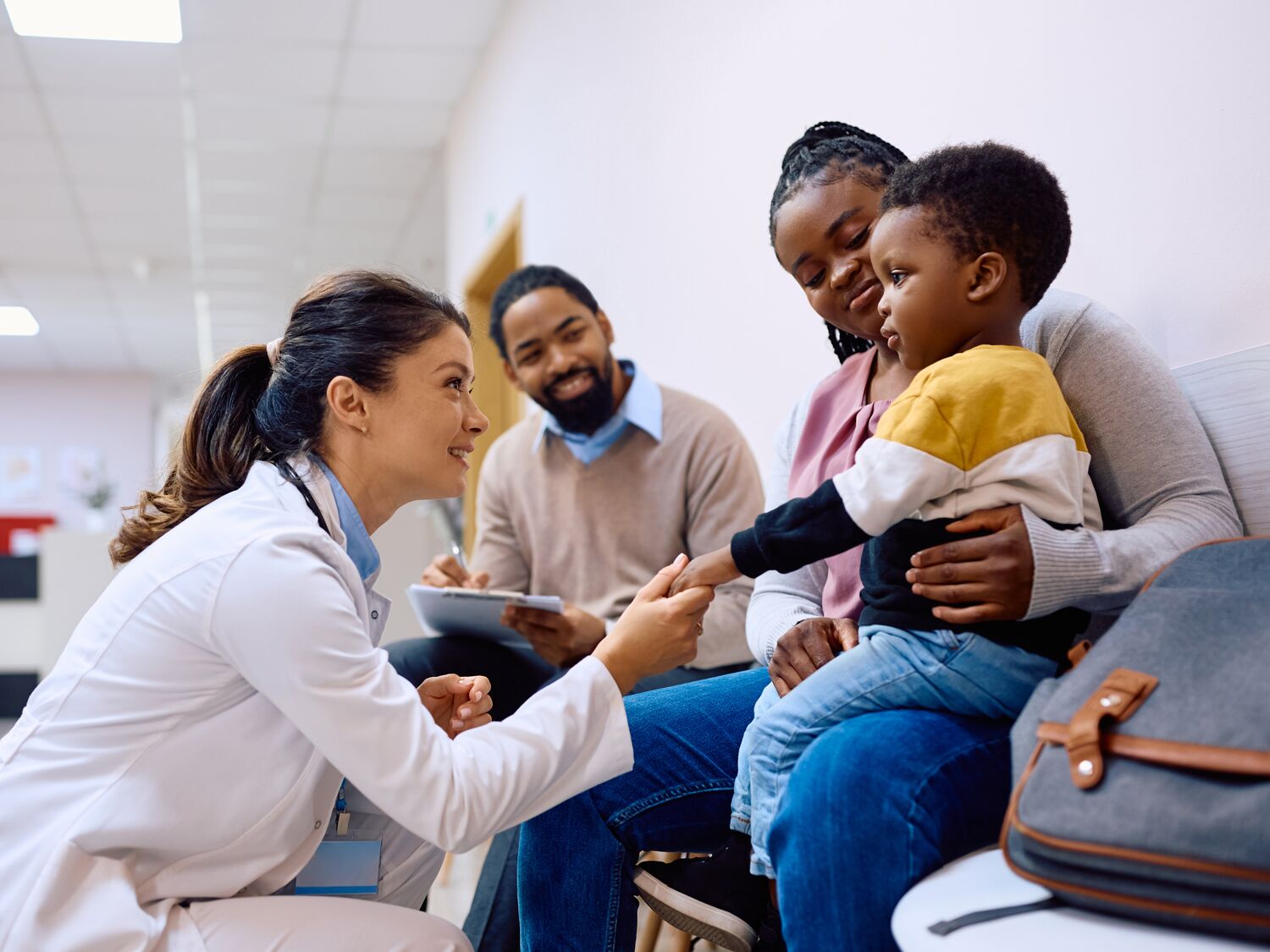 Smiling doctor holding hand on African American boy who came with parents for medical exam at pediatric clinic.