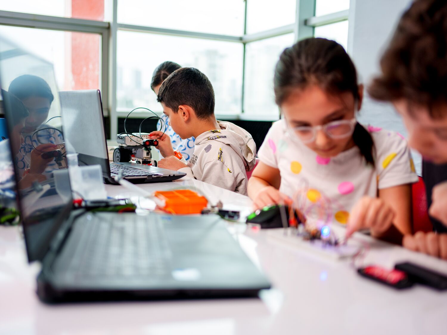 Group of young students participating in a hands-on robotics and STEM education activity. Two children are assembling a robot with various components, while others work on coding and programming using laptops. The classroom is vibrant and filled with educational tools and materials, promoting creativity, teamwork, and innovation. Perfect for themes related to technology, education, learning, and future skills development.