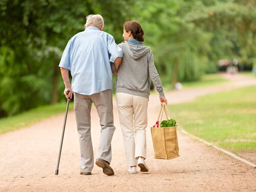 An elderly man walking with his caretaker who is holding a grocery tote.