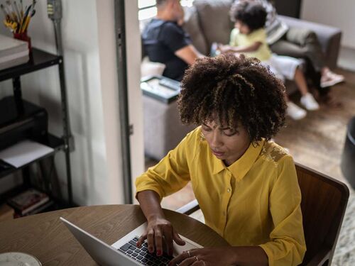 Woman working on our laptop at home, her family on the couch behind her