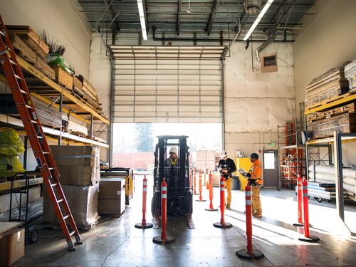 A multiracial woman worker operates a forklift, her coworkers reviewing paperwork for the day in a warehouse setting, wearing safety gear.