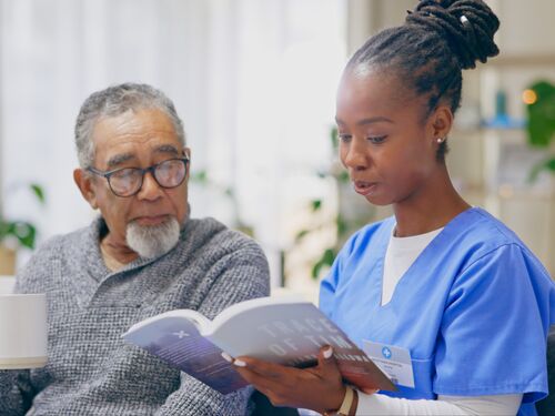 A nurse is reading a book next to a an old patient who is holding a cup and looking over at the book.