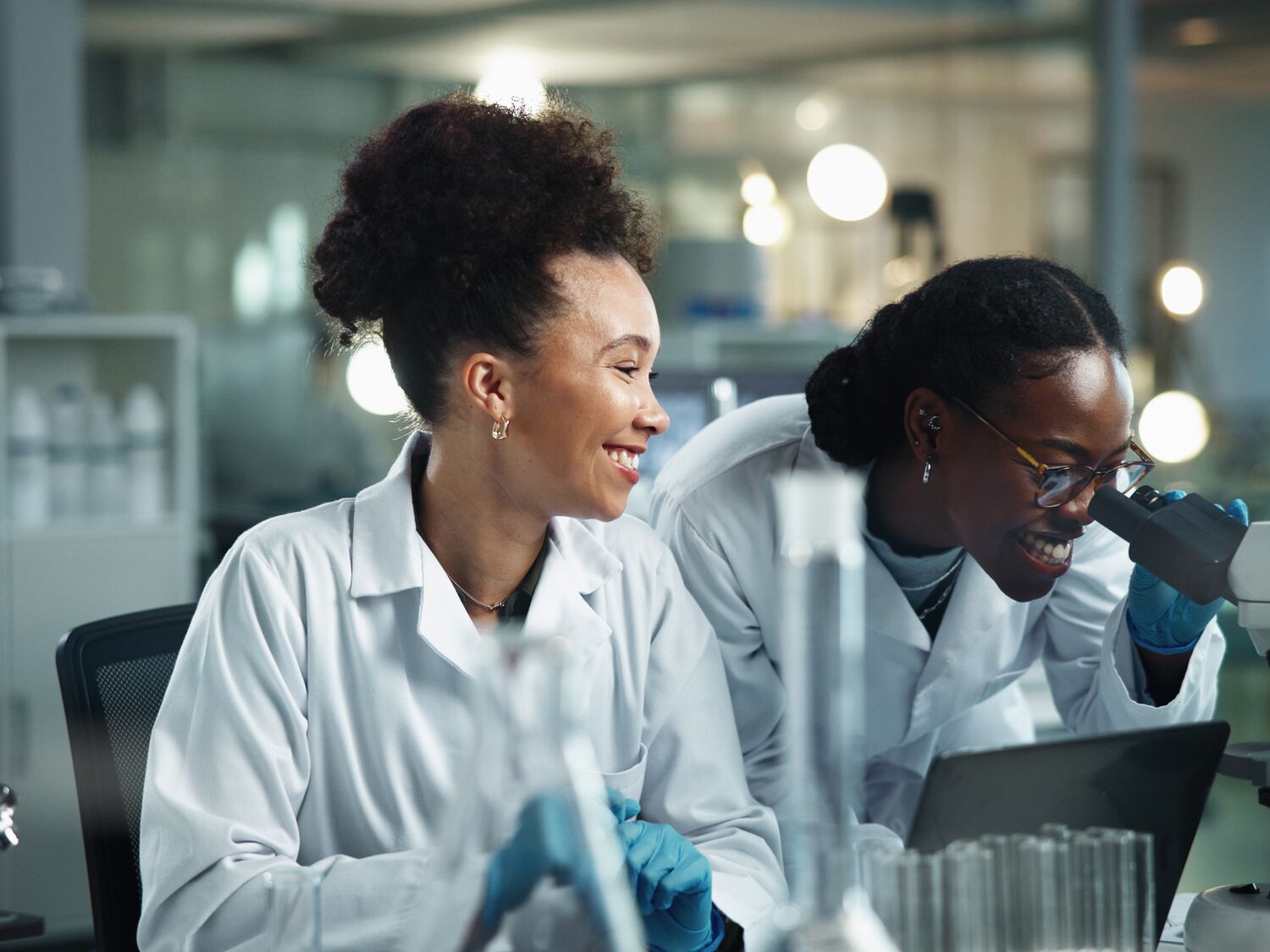 One scientist is looking inside a microscope while another scientist is sitting next to her.