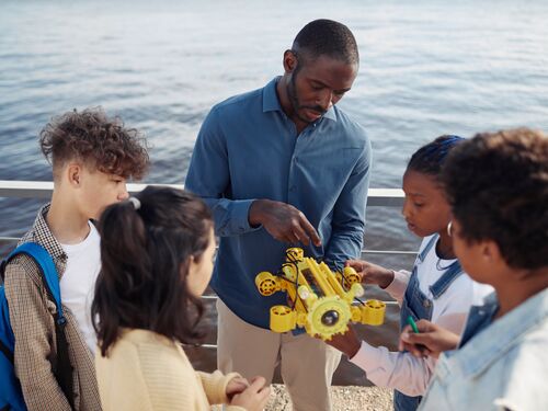 Portrait of black male teacher demonstrating robot model to group of children standing in circle during engineering class outdoors