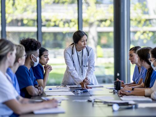 A small group of medical student residents gather around a boardroom table to to meet with their medical team lead.  They each have cases out on front of them as they work together collaboratively to discuss each one.  They are each dressed professionally in medical scrubs and are listening attentively to the doctor leading the meeting at the head of the table.