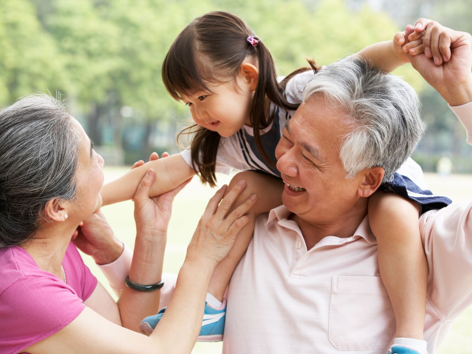 A young girl sits on her grandfather’s shoulders while her grandmother reaches up to hold her hand in a park.