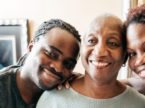 Three people smiling warmly, with an older woman in the center and two younger adults leaning in close on either side in a cozy indoor setting.
