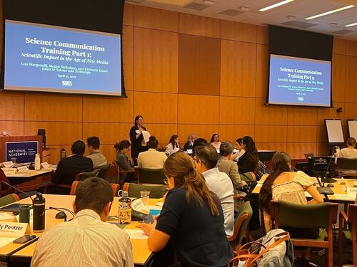 Moderator standing at table speaking to a group of collaborators.