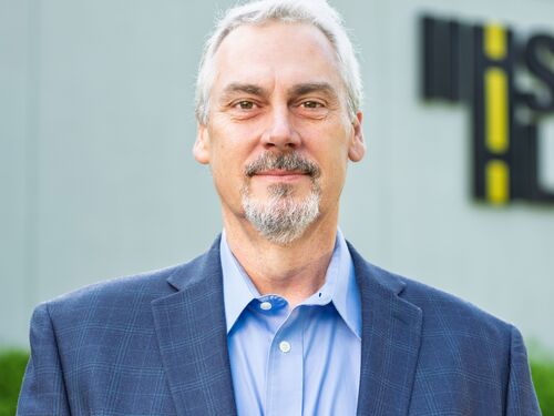 A white man with white hair and goatee standing in front of a grey background with a blue shirt and dark blue sportscoat