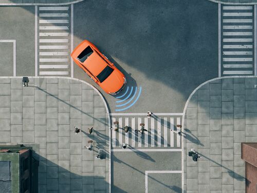 Aerial view of a bright orange car driving on a street. The car is next to a pedestrian crossing and in front of a stop line with the word "Stop" painted on the road. The car uses sensors and radar to detect the pedestrians. Concept of new car technology like autonomous driving.
