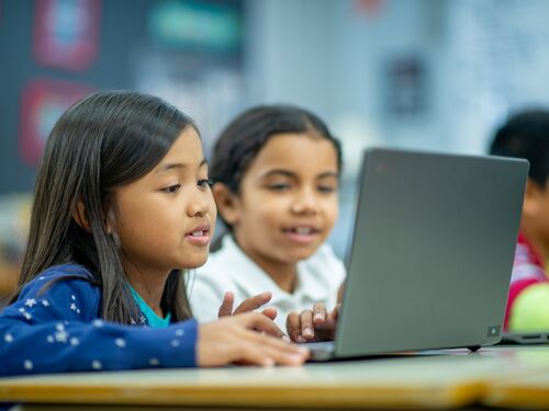 Two Elementary students are seen sitting side-by-side with a laptop open in front of them during a computer lab.  They are each dressed casually and focused on working online together.