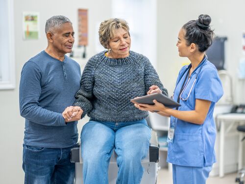 A patient sits up on an exam table with her partner close by as she receives her recent tests results from a doctor.