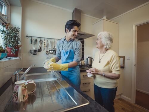 Teenage boy is talking to his grandmother while he washes the dishes in her kitchen.