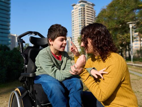 Cheerful child sitting in a wheelchair communicating with his mother using sign language in a park