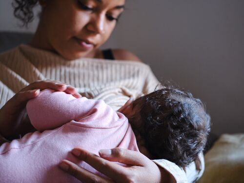front view portrait of a confident Black mother looking tender her little baby in her arms while breastfeeding sitting in the sofa during a regular day at home. concept family and motherhood